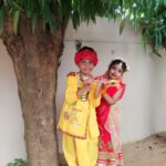 Two children in festive indian costumes pose under a tree boy in yellow krishna attire with a peacock feather headpiece holds a flute girl in red gesture dress stands beside him as radha with floral headpiece